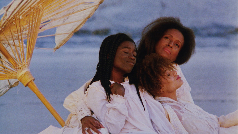 Three black women, all with white clothing, sitting and embracing under a wooden umbrellla at the beach