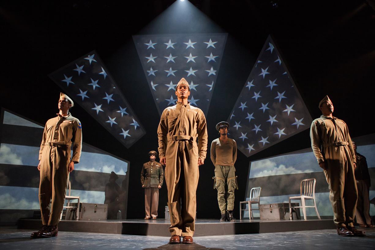 photo pf the play being formed. There are 4 men in uniform and military fatigues spaced out on the stage, facing the audience, and standing at attention.