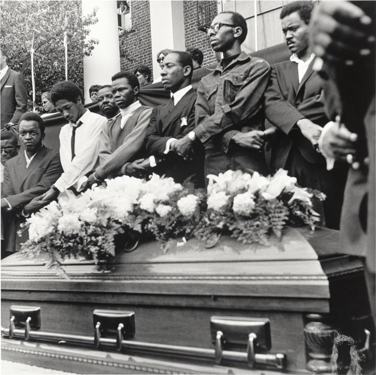 photo showing six black men standing in front of a building and behind a coffin holding hands with crossed arms .
