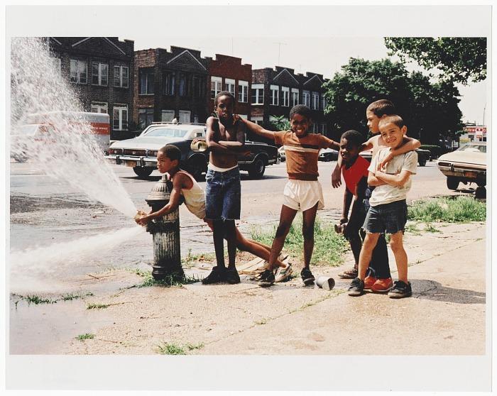 This color photograph shows young boys opening a fire hydrant on a summer day in the Brownsville section of Brooklyn, New York, in 1980