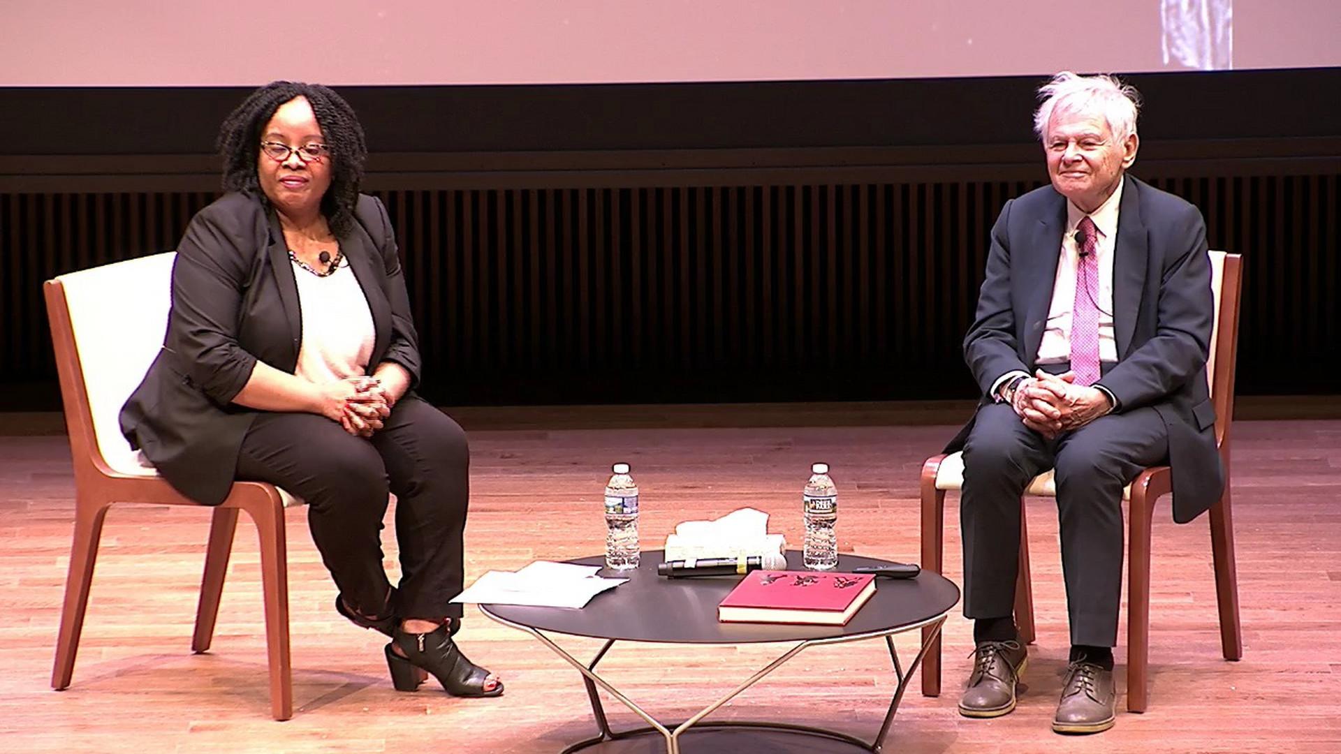 photo of an black woman and a white man sitting in chairs on a stage with a low table in between them.