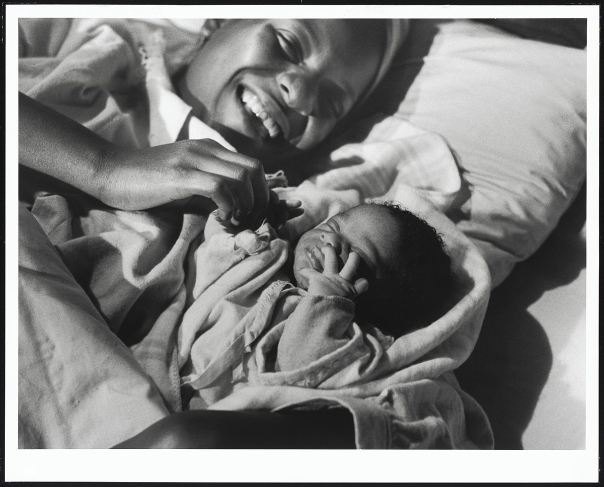 Black and white image of Martha Sapp holding a baby, both lying in bed. Sapp lays in a bed, covered in blankets and sheets, smiling, open-mouthed. Her proper right hand holds the proper left hand of her child. Her head rest on a pillow. She and the baby take up most of the image, diagonally, from the upper left to the lower right. the baby lays eyes closed and hands raised to its face. The child is wrapped in a striped, plaid blanket.