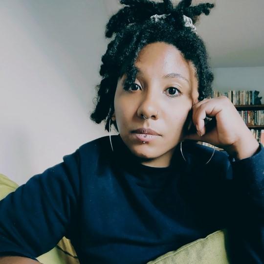 African American woman wearing a black shirt stares at the camera. Her head rests against her left hand with fingers at her temple.