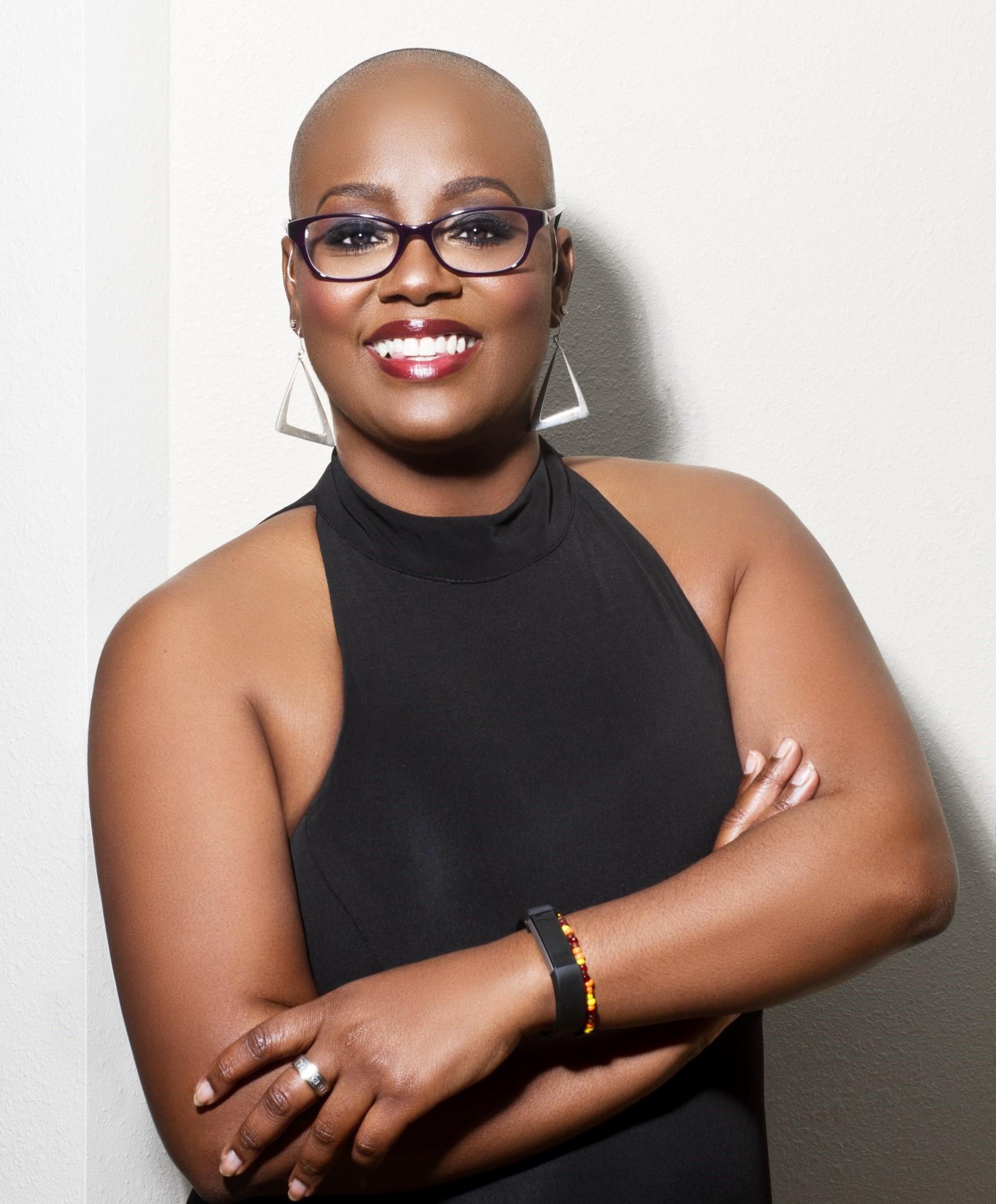 African American woman with a bald head smiles to the camera. She wears a black halter top and her arms are folded across her front body. She wears glasses with black frames.