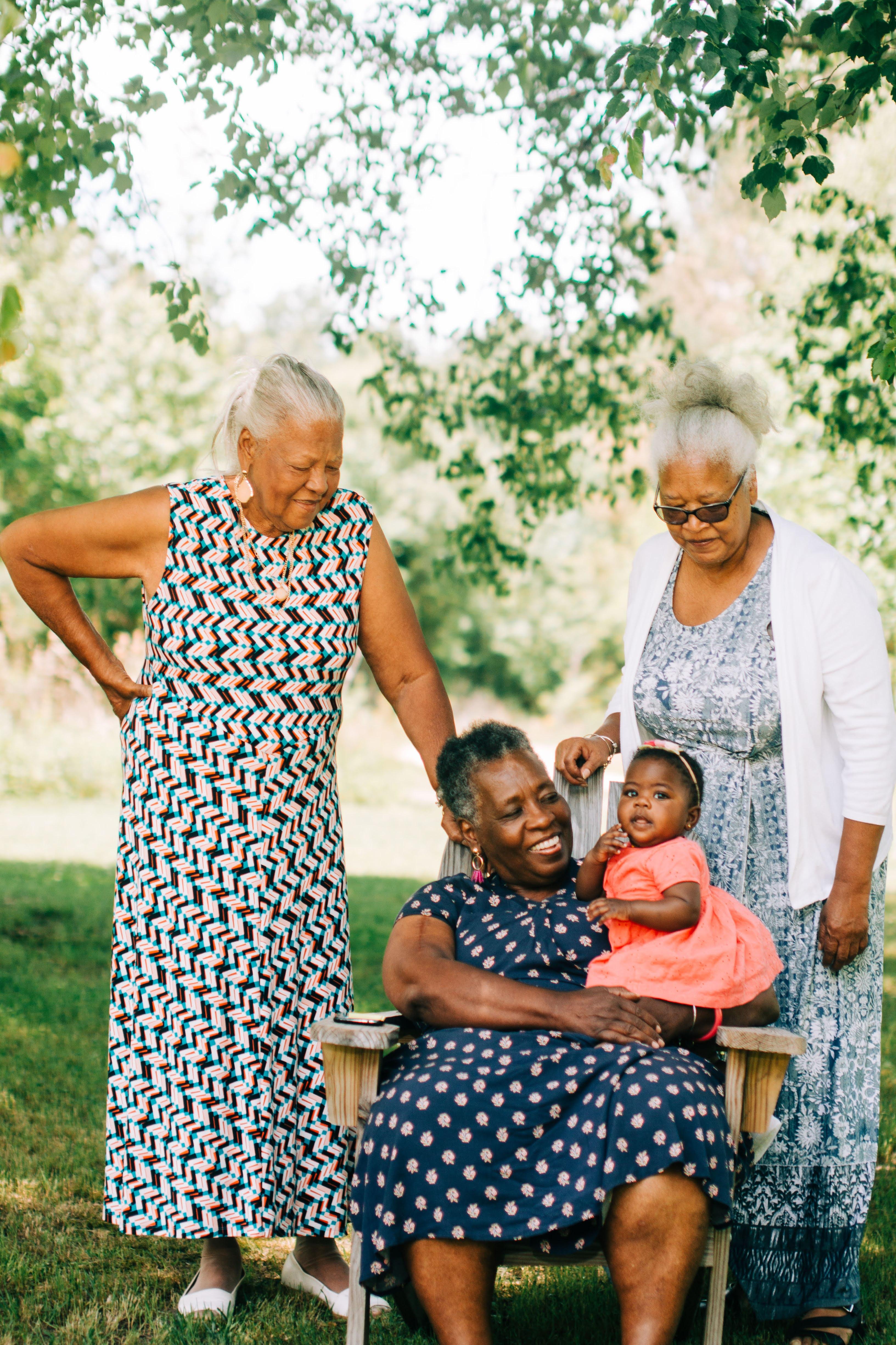 The older black women smiling at a baby. Two women are standing, and the third is seated and holding the baby.