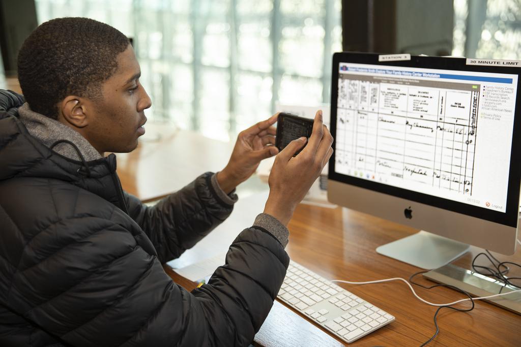 A young man points a cell phone at a computer in the Family History Center