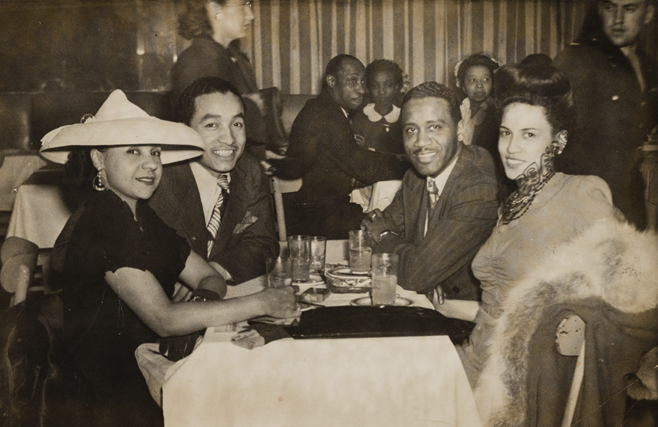 Four people seated at a table covered with a white table cloth, small plates, and glasses.