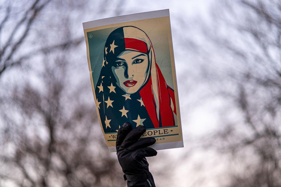 A man holding up a poster which shows a Muslim woman wearing a hijab with an American flag design with the text around her reading, “We the People Are Greater Than Fear.”