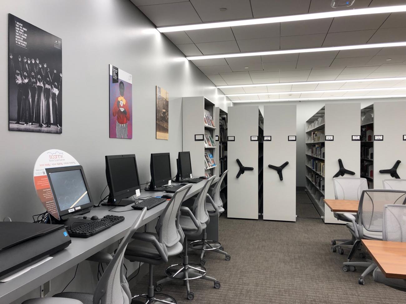 Room with a narrow table against the wall on the left with 4 computer screens and desk chairs. In the back of the room our several movable stacks storing the library collection. On the right are two tables.