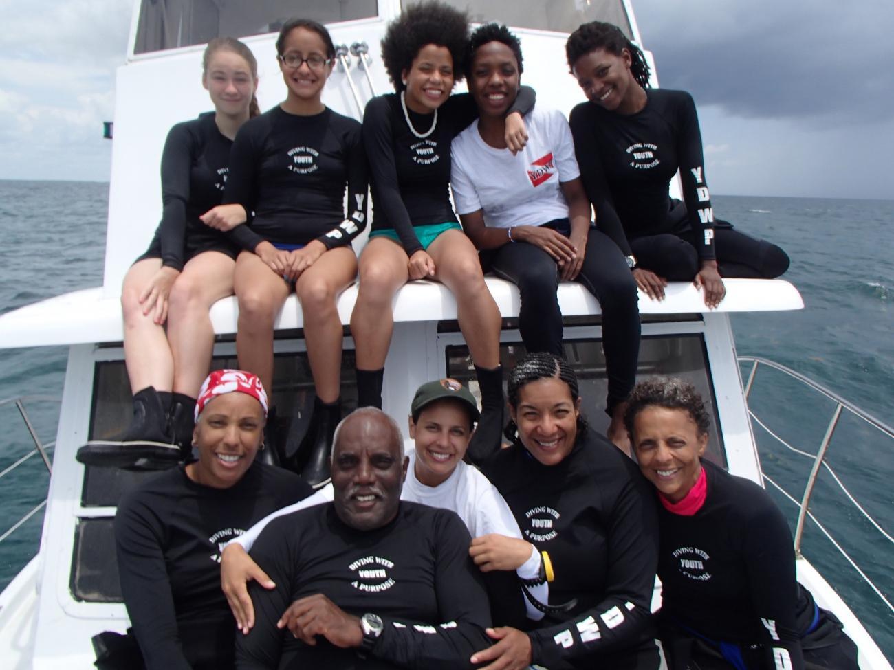 A group of ten scuba divers in wetsuits sit on a boat in the ocean.