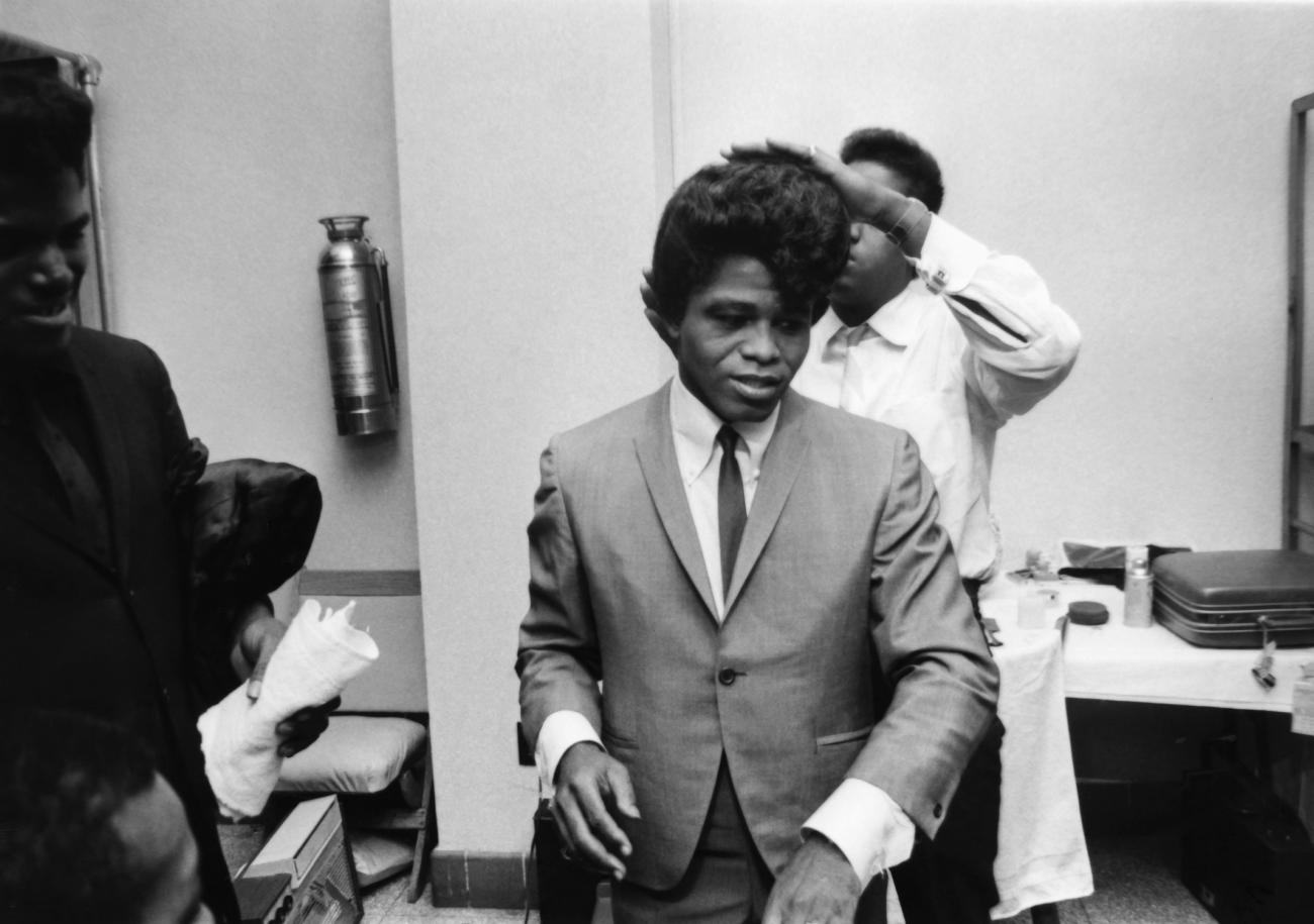 Black and white image of James Brown combing his hair in a grey suit and black tie.