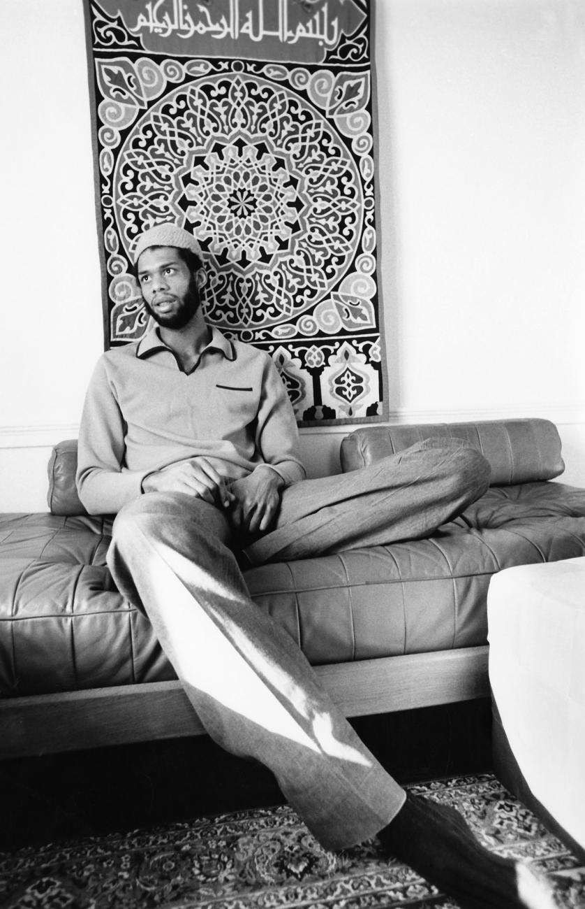 Image of Kareem Abdul Jabbar sitting in front of a muslim prayer rug on a bench