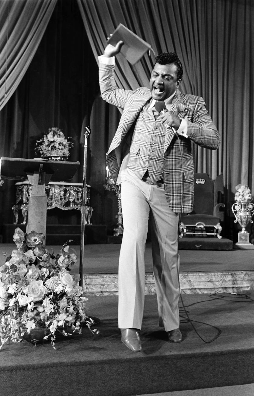 Image of Reverend Ike in a pulpit with aBible in his hand as if he might throw it.