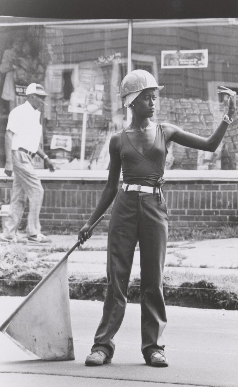 A female construction worker wearing a hard hat and holding a flag in her right hand