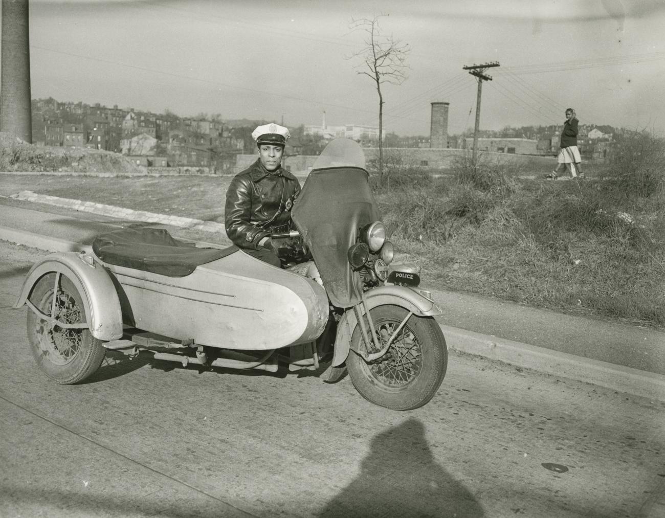 A black and white photograph of a policeman sitting in a motorcycle with a sidecar. 