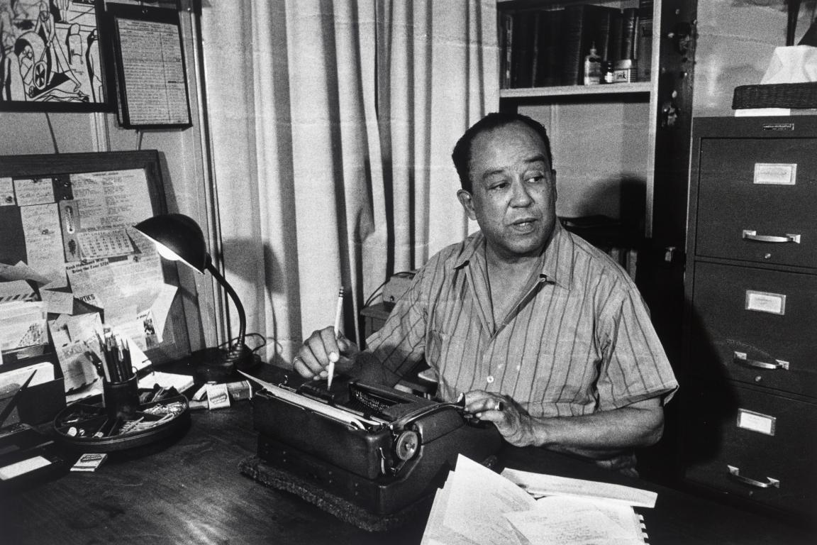 Langston Hughes at work in his office. He holds a pencil as he sits in front of his typewriter at his desk.