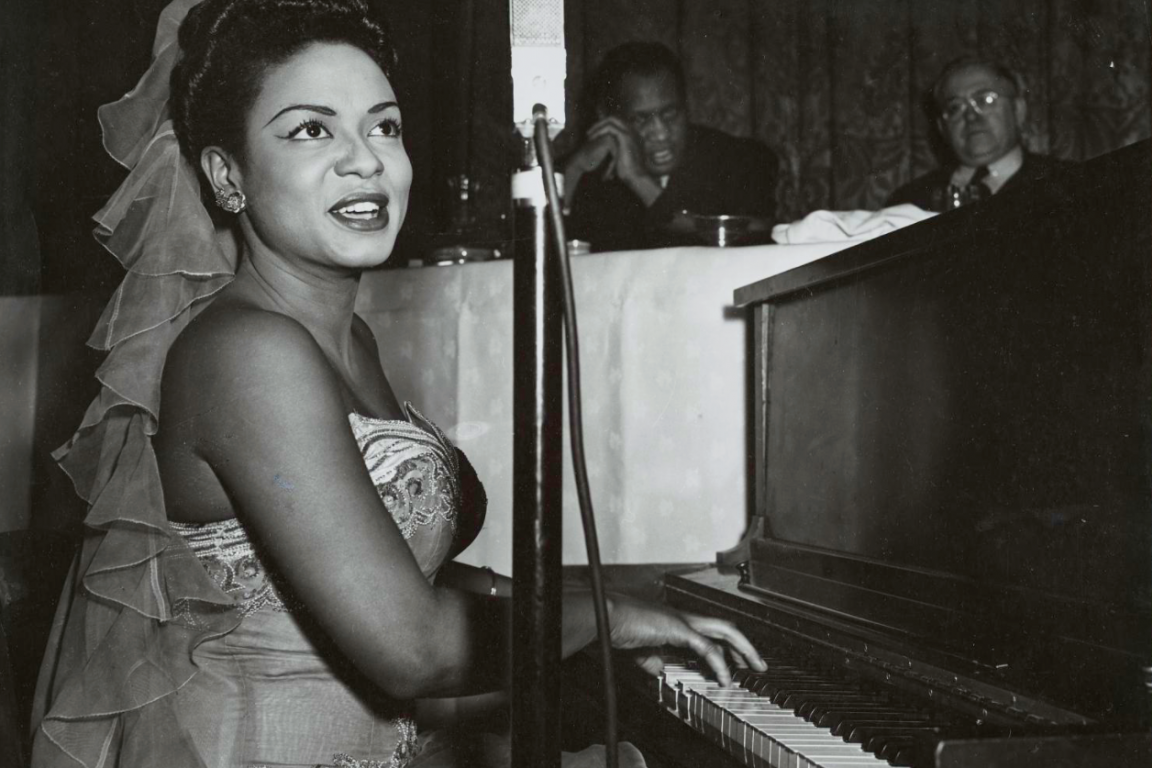 A black-and-white photograph of Hazel Scott and Paul Robeson in a New York City club. Scott plays the piano and sings into a microphone.