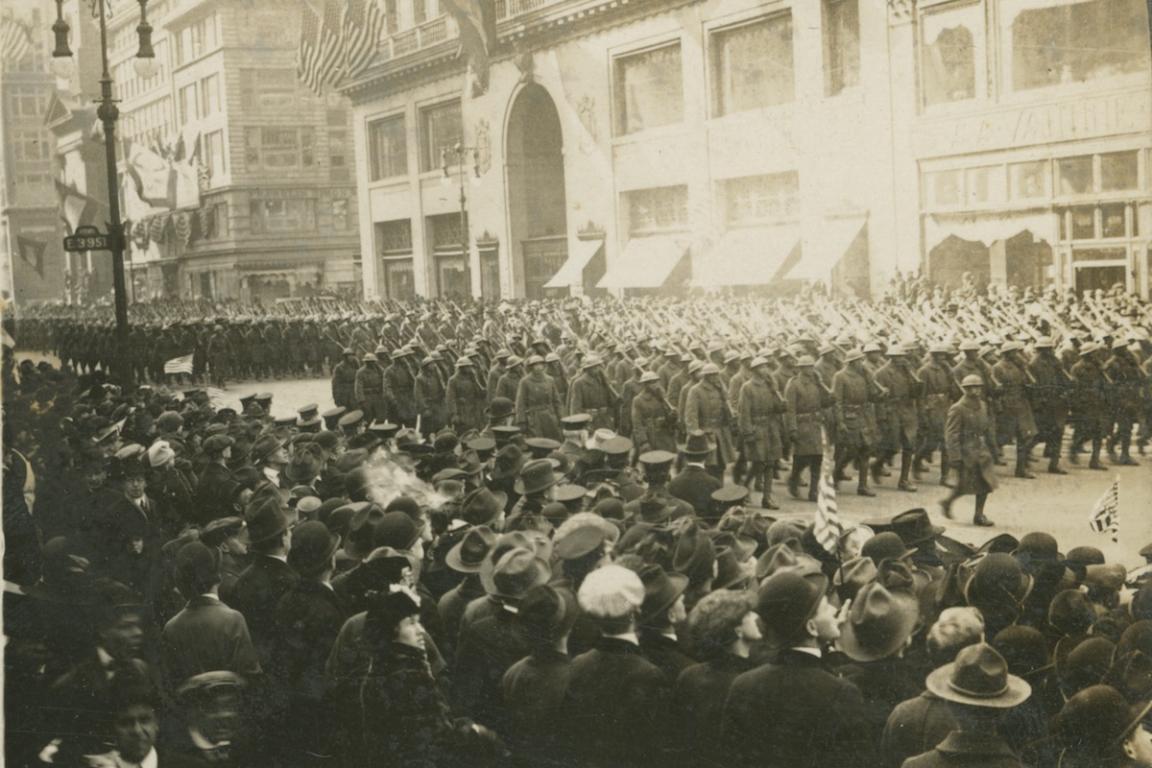 Black and white photo of soldiers in military formation marching down a city street. Crowds line the route with tall buildings in the background.