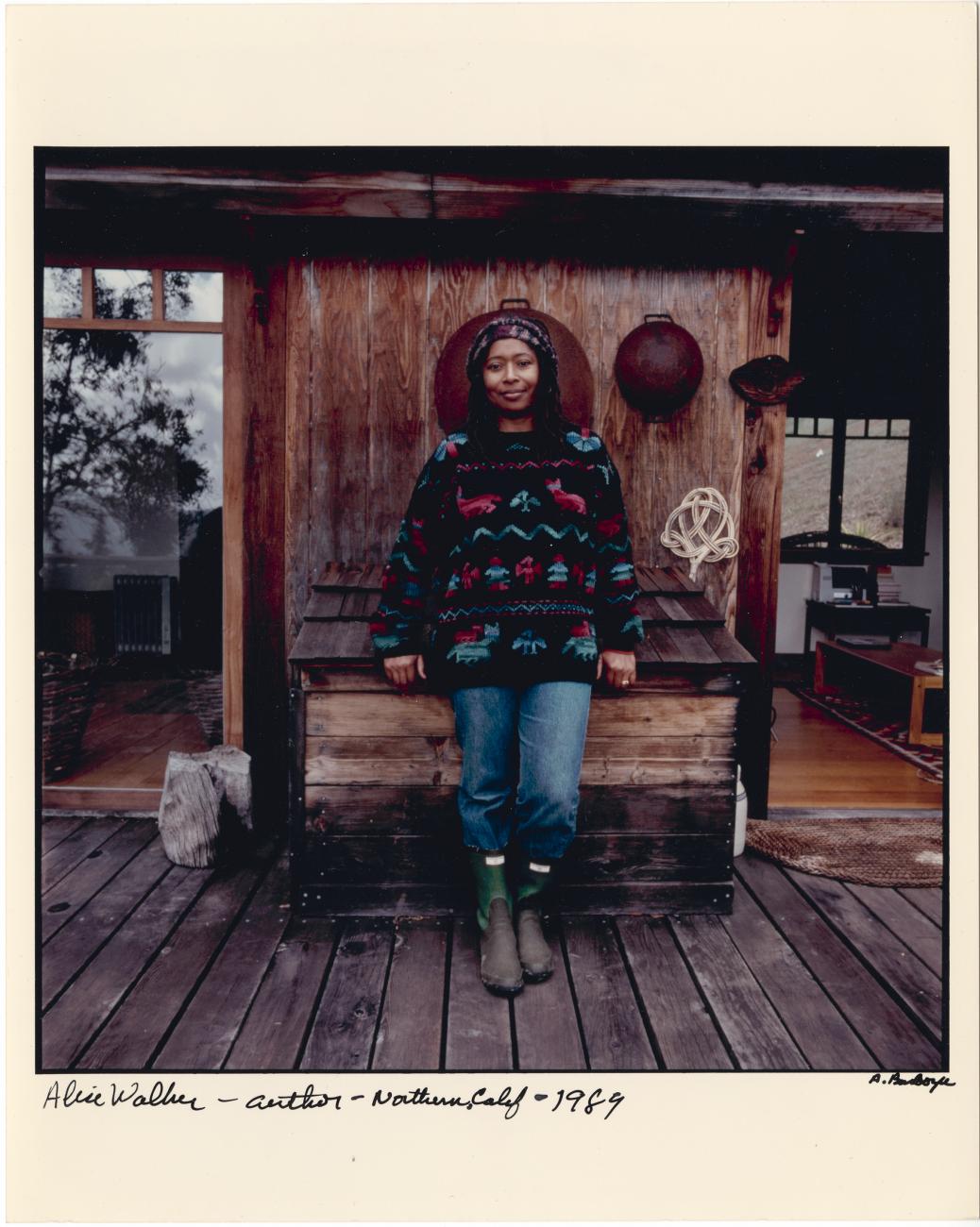 Color photograph of Alice Walker leaning against a wooden bin on the porch of a house. 