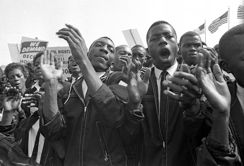 A digital image of The March on Washington for Jobs and Freedom, which took place in Washington, DC on August 28, 1963. The image depicts a close-up of a crowd of men and women clapping and chanting or singing. In the background, placards and American Flags are visible.