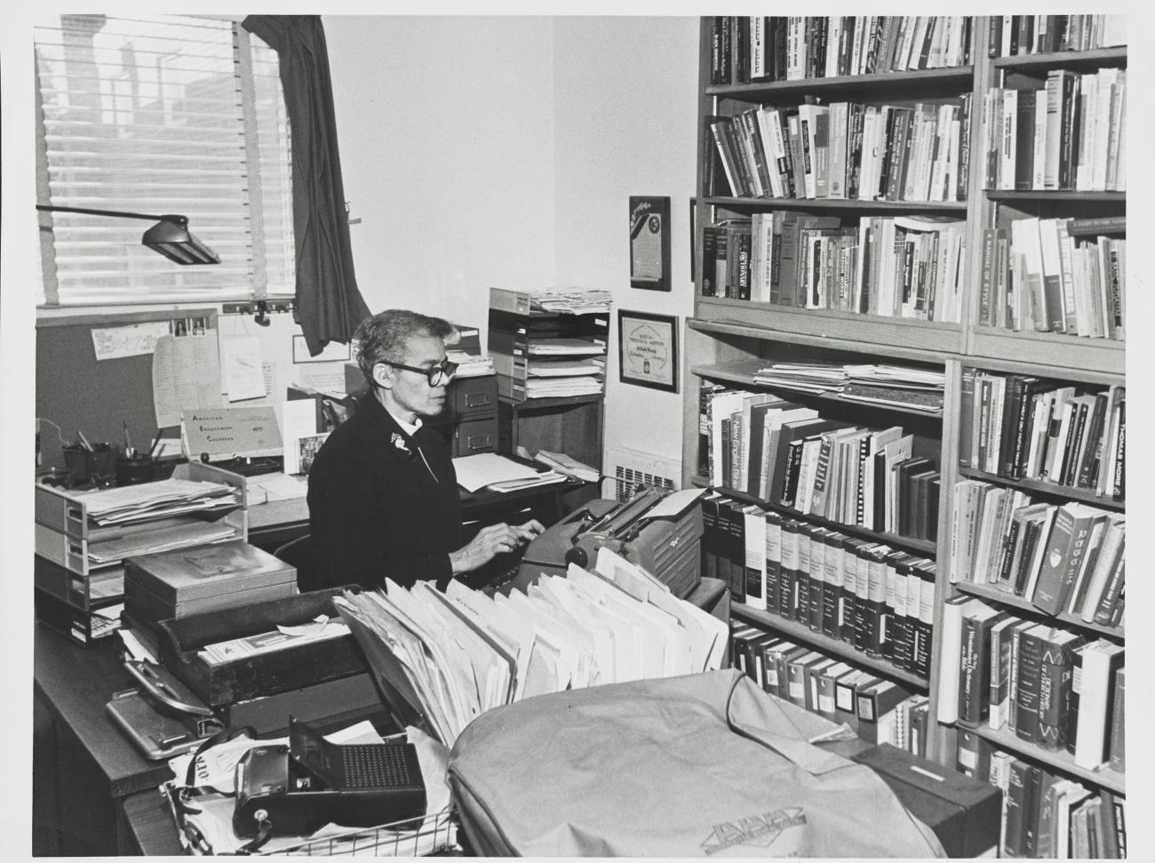 Pauli Murray sitting at a typewriter by a desk crowded with papers.