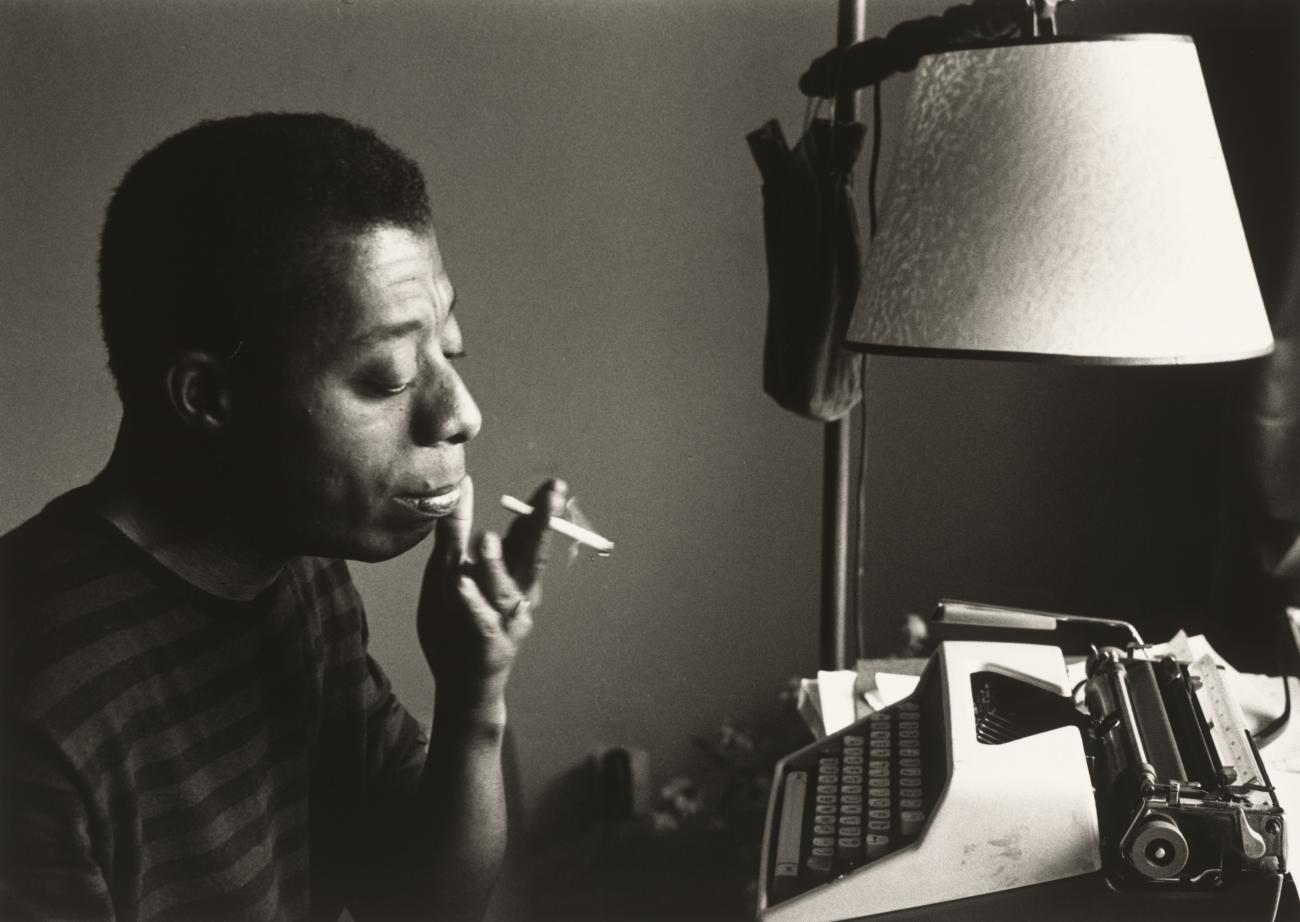Image of James Baldwin smoking a cigarette sitting at a typewriter.