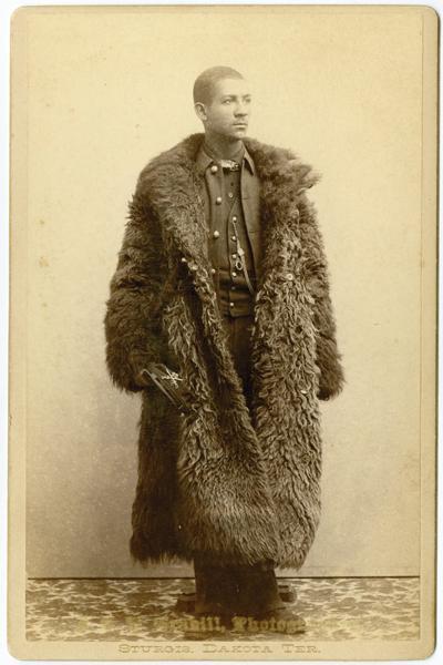 Photograph shows a standing black soldier wearing an unbuttoned buffalo hide overcoat.