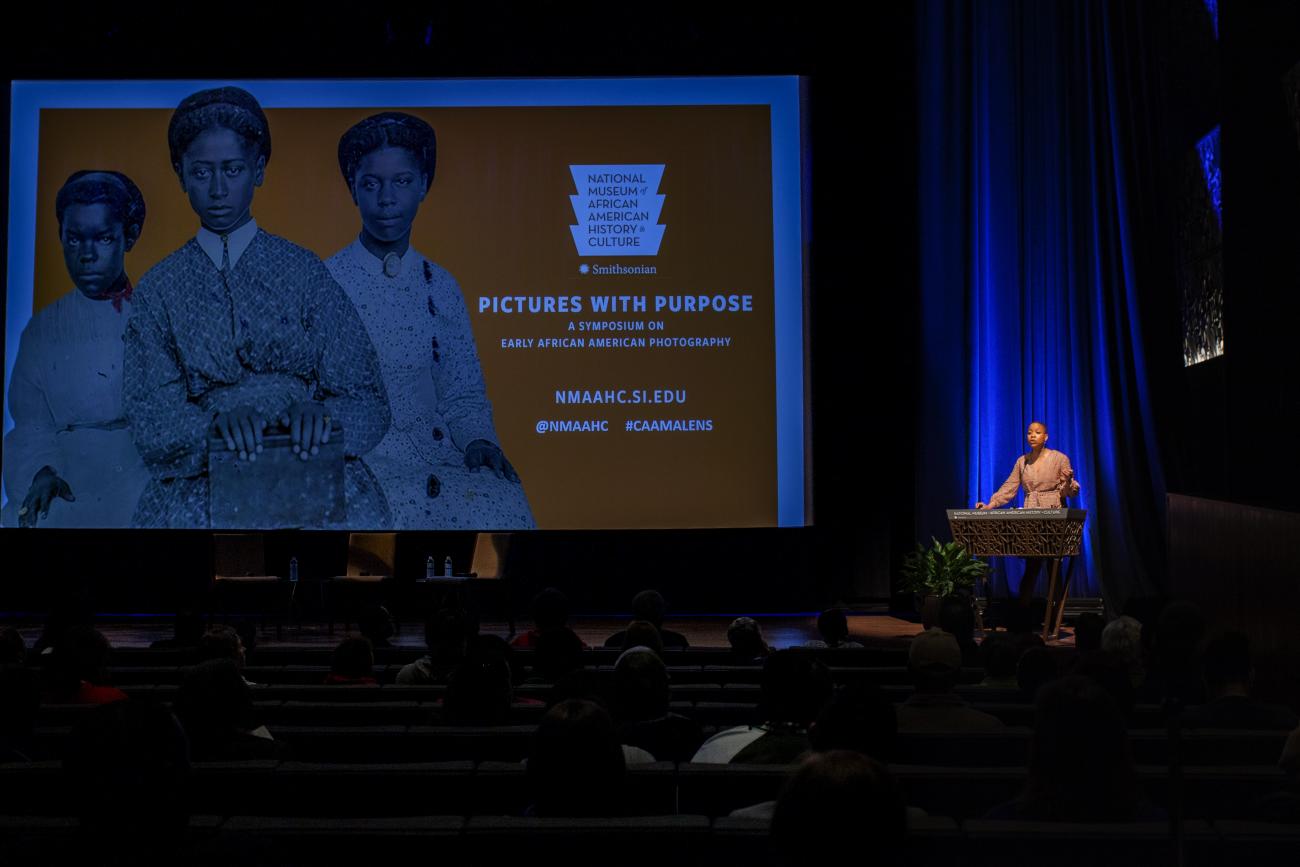 A color photo of a woman standing on stage in front of a podium talking into a microphone. A logo with three women in old fashion clothing and an orange background is projected onto the screen on stage.