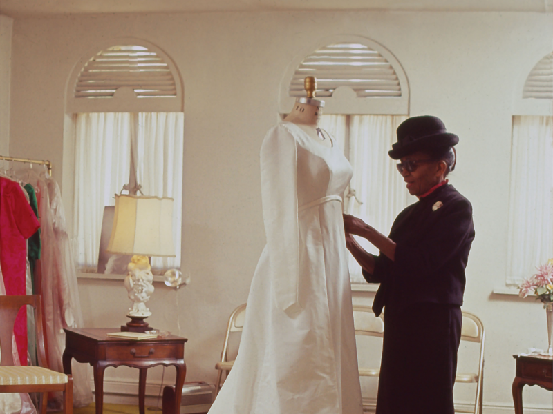 image of anne lowe standing next to a mannequin. She has on a black skirt and jacket. The mannequin is dressed in a white gown.