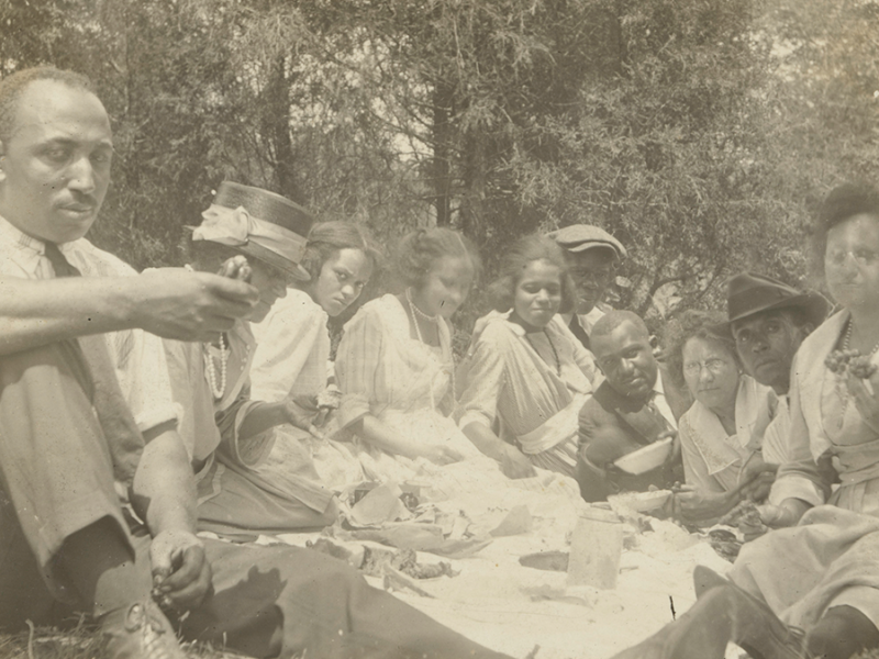 Black and white image of a black family at a picnic. Family is seated in a semi-circle around a blanket, facing the camera.