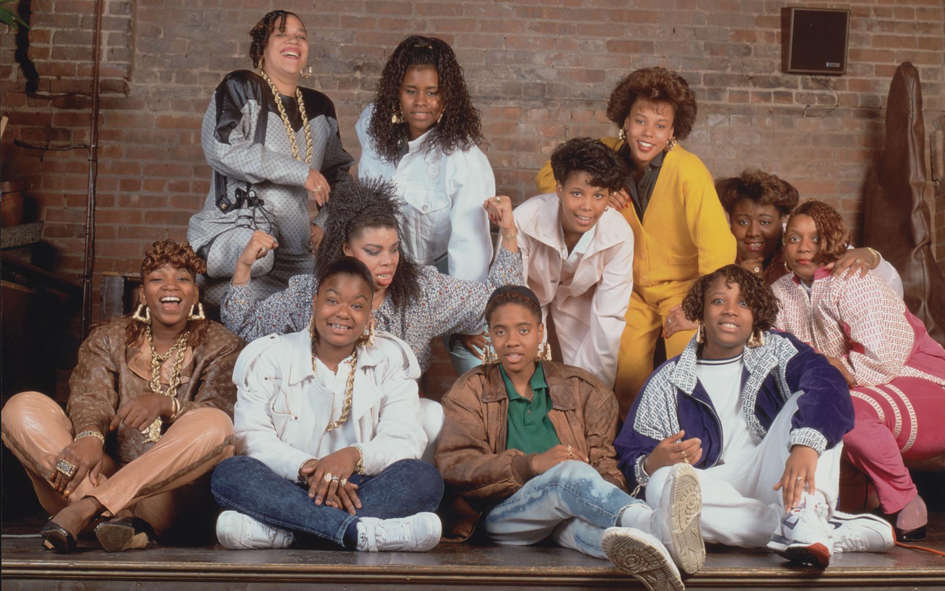 A color photograph of eleven female rappers posed together on a wooden stage in front of a brick wall. 