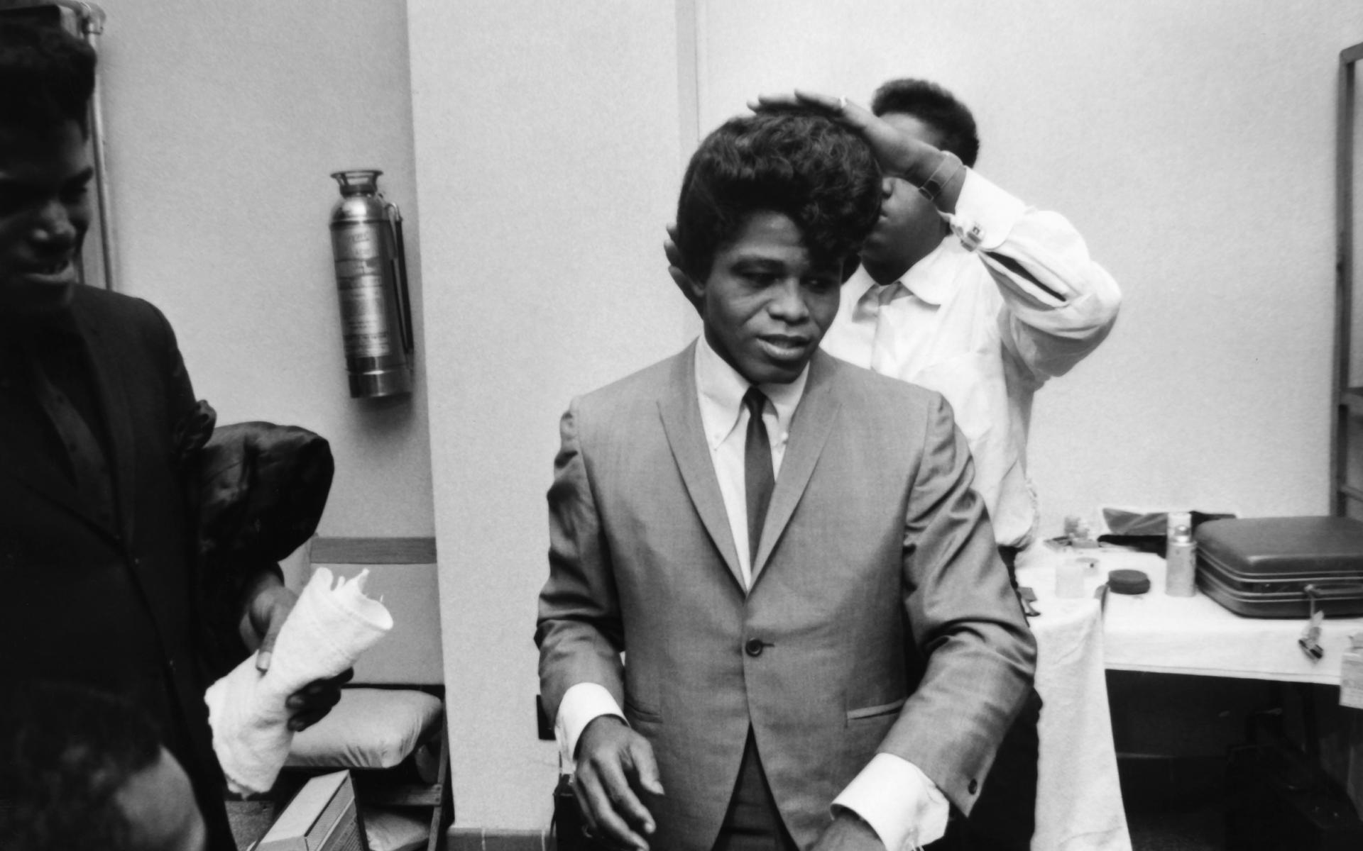Black and white image of James Brown combing his hair in a grey suit and black tie.