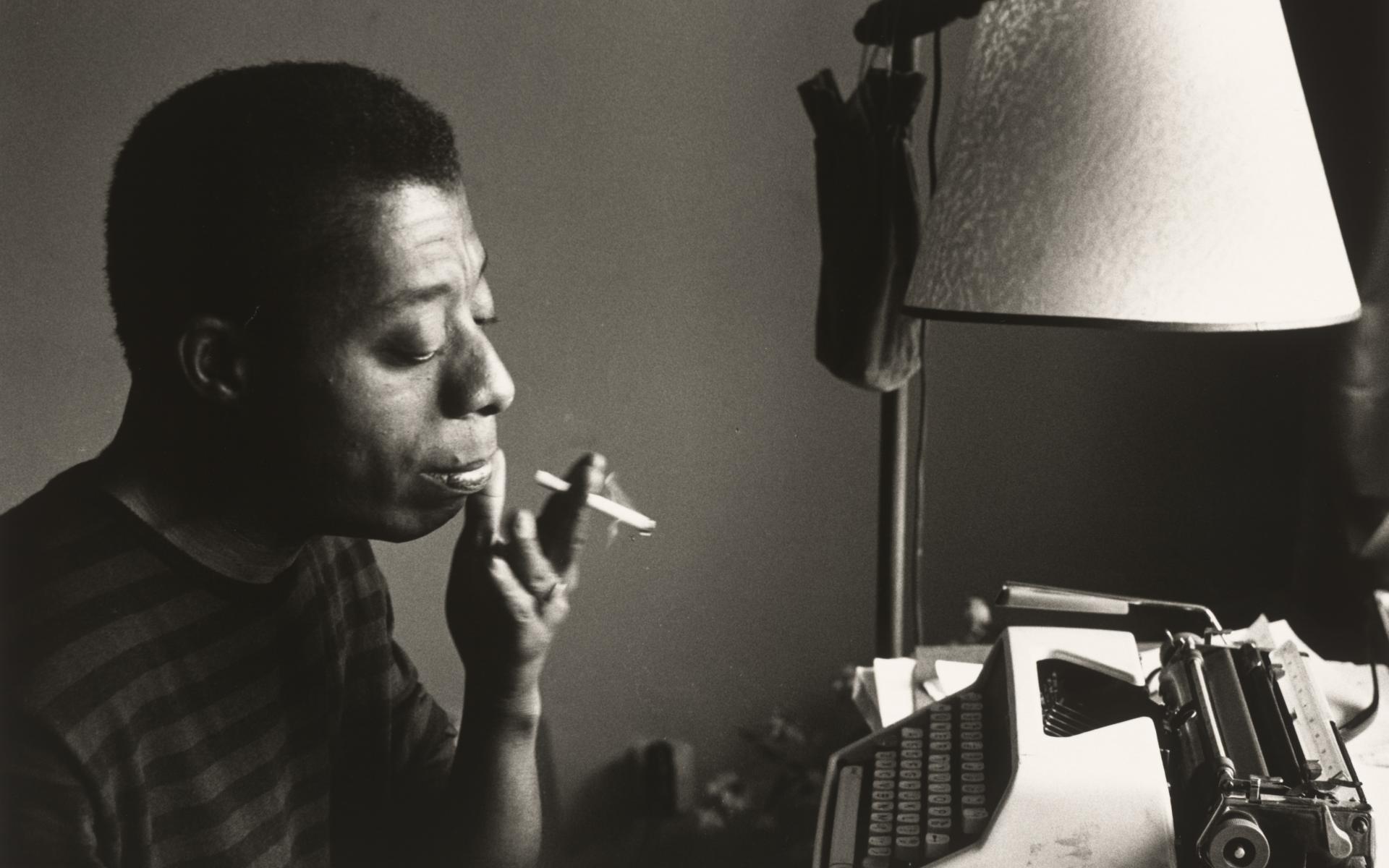 James Baldwin smoking a cigarette while sitting at a typewriter. 