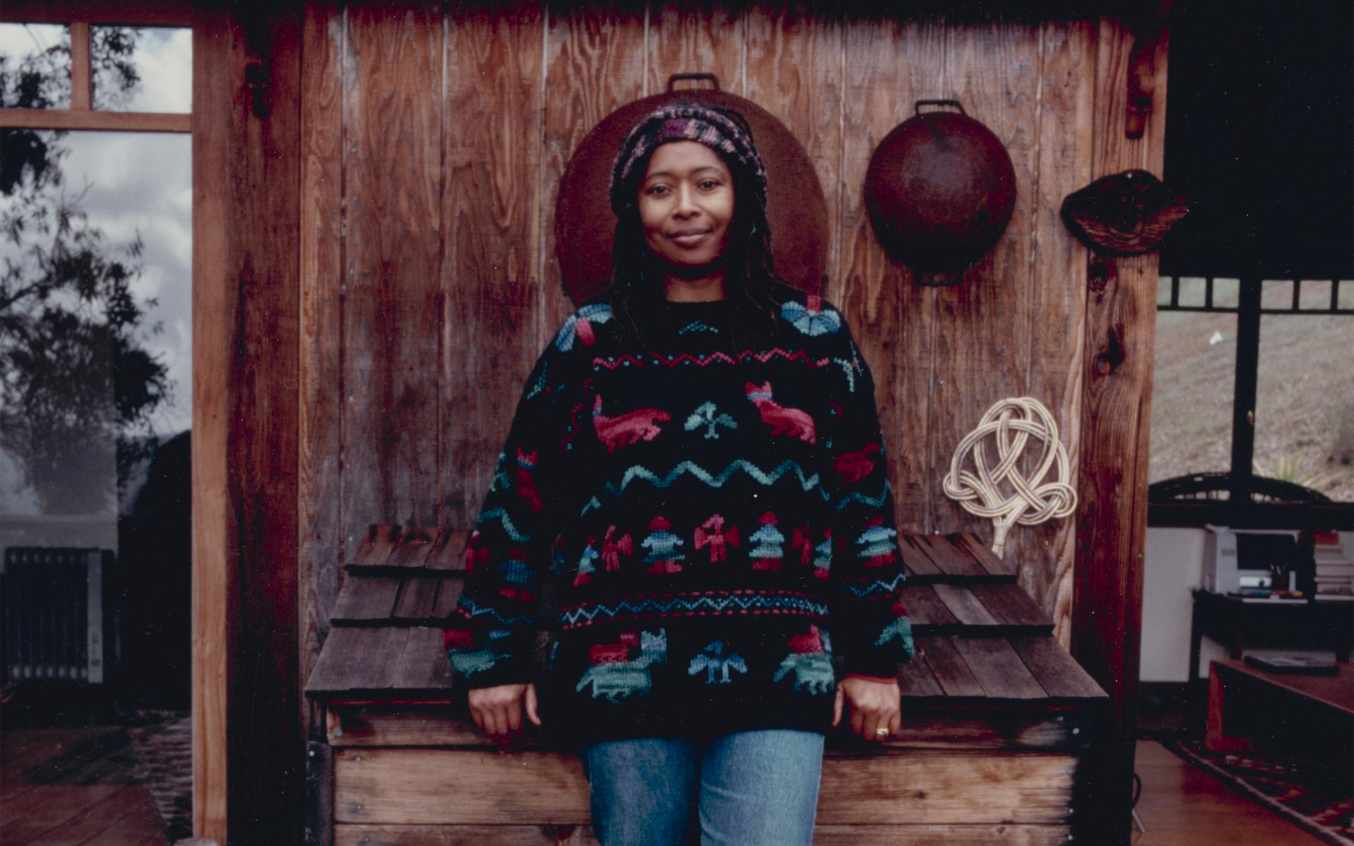 Color photograph of Alice Walker leaning against a wooden bin on the porch of a house. 