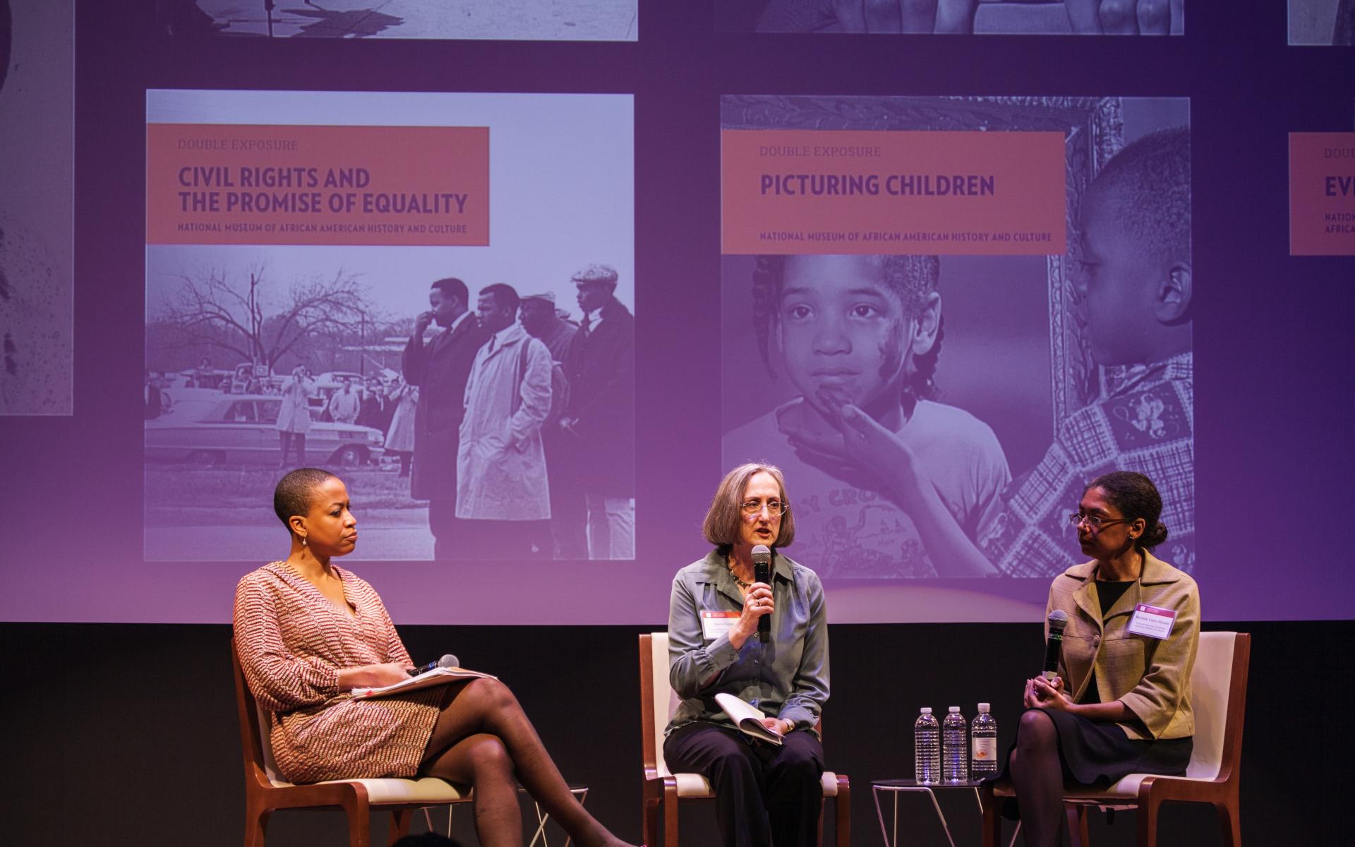 A color photo of three women sitting on stage and holding microphones. The woman in the middle and the other two women are looking at her. Behind them the covers of several books are projected onto a screen.