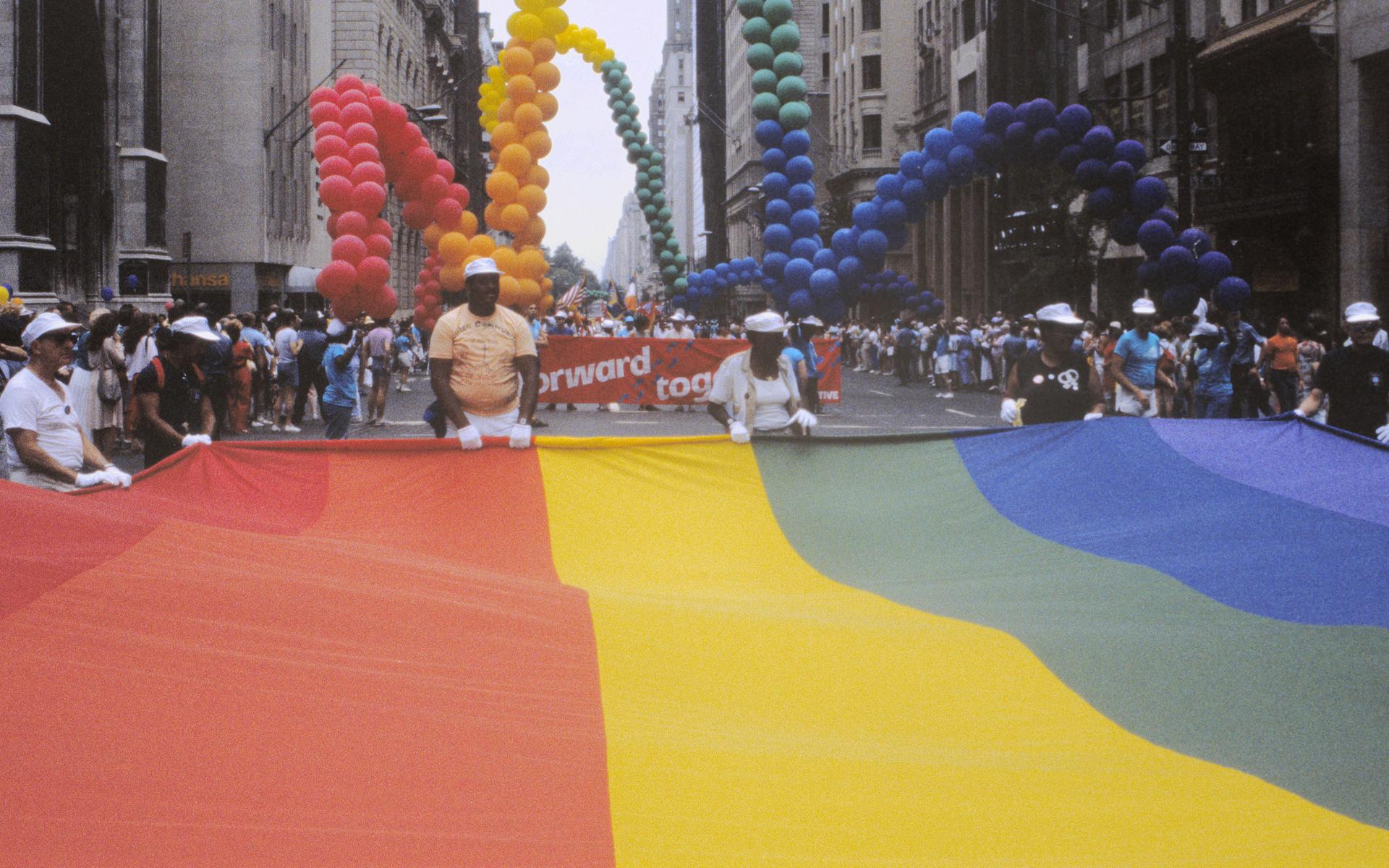 Image of men holding a large rainbow flag with buildings and balloons in the background.